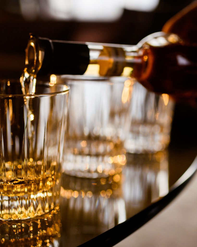 Close up of anonymous man pouring gold whiskey from bottle in glass which standing on wooden table indoors